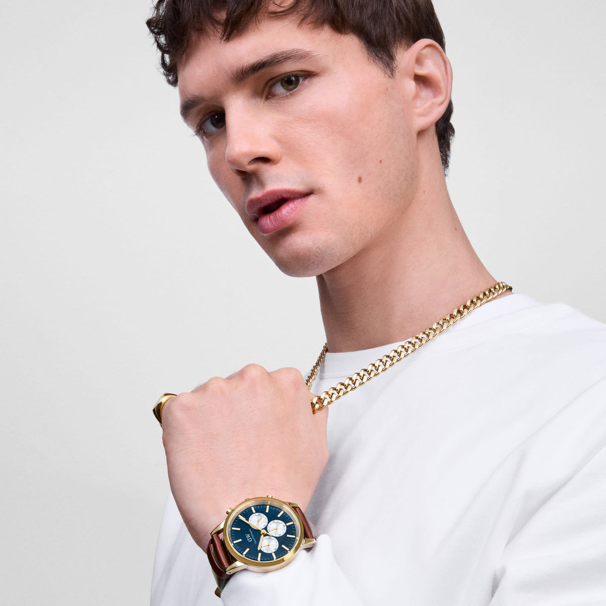 Male model wearing the Daniel Wellington Iconic Chronograph watch, featuring a gold case, an arctic blue panda dial, and a brown St Mawes leather strap.
