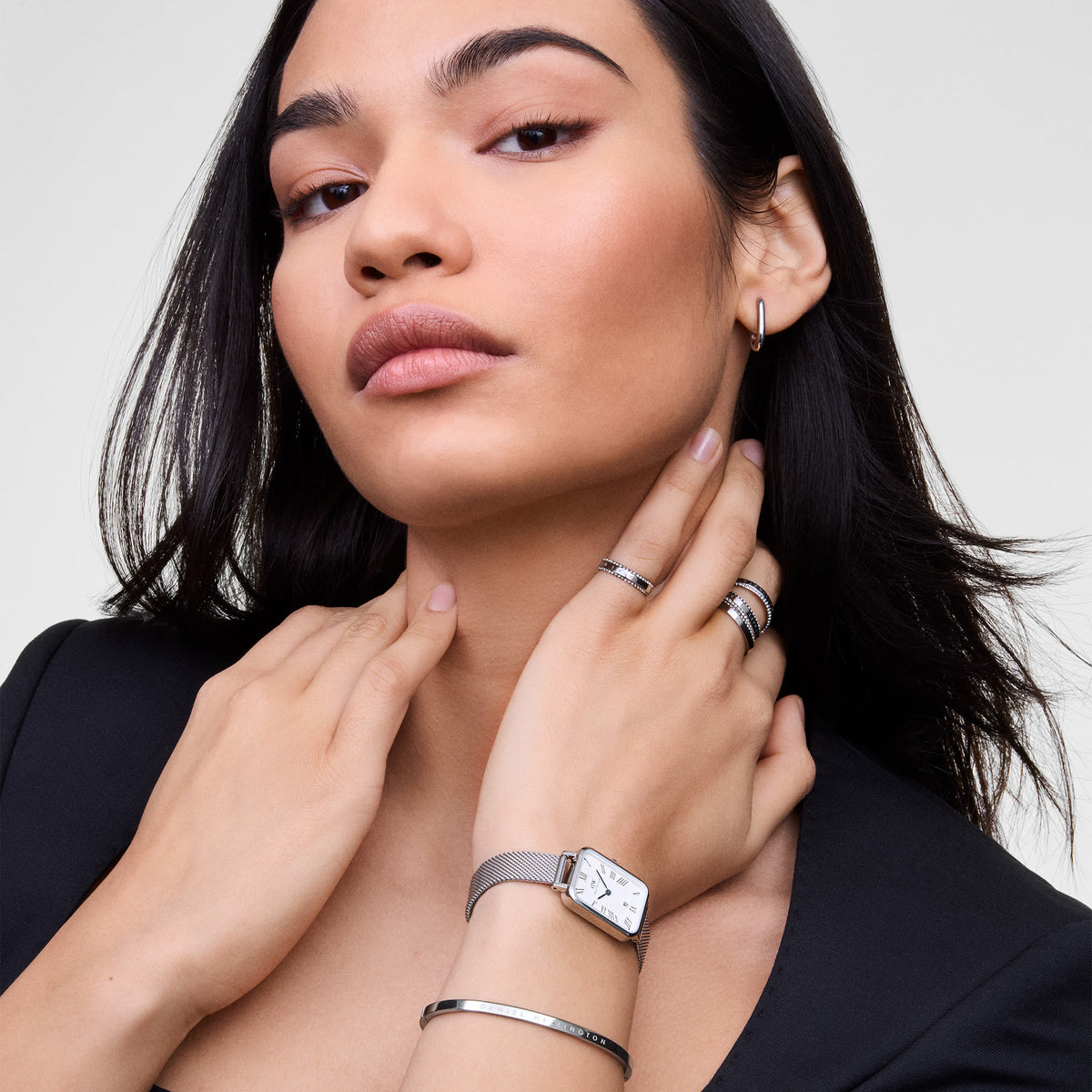 A woman with dark hair wears the Daniel Wellington Quadro Sterling watch, featuring a rectangular white dial with Roman numerals and a silver mesh strap.