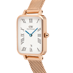 Daniel Wellington Quadro Melrose watch in rose gold with a rectangular white dial, Roman numerals, blue hands, a date window, and a mesh strap.