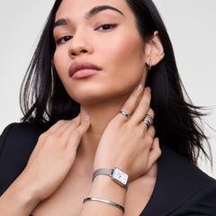 A woman with dark hair wears the Daniel Wellington Quadro Sterling watch, featuring a rectangular white dial with Roman numerals and a silver mesh strap.